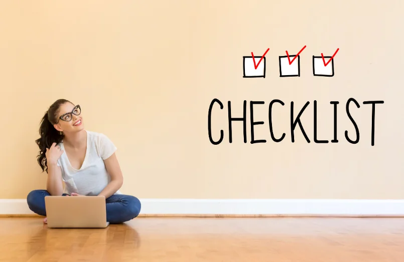 Woman thinking at laptop, checklist on the wall